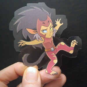 May include: A clear sticker featuring a cartoon character with red, yellow, and brown hair, wearing a red and white outfit with a yellow belt and a red cape. The character has a fierce expression and is in a dynamic pose with one leg raised.