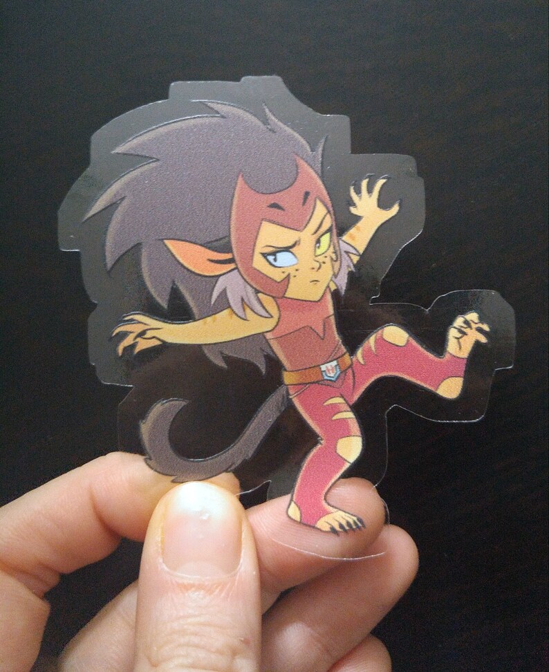 May include: A clear sticker featuring a cartoon character with red, yellow, and brown hair, wearing a red and white outfit with a yellow belt and a red cape. The character has a fierce expression and is in a dynamic pose with one leg raised.