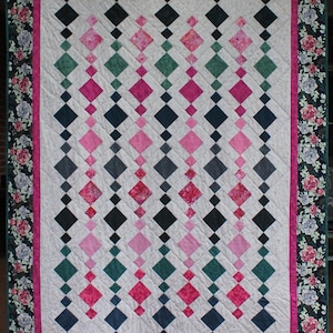 Op de afbeelding: Een handgemaakte quilt met een witte achtergrond en rijen afwisselende ruitvormen in roze, groen, grijs en marineblauw. De quilt is omrand met een bloemenpatroon in roze, groen en wit.