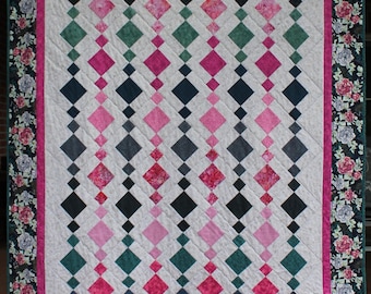 Op de afbeelding: Een handgemaakte quilt met een witte achtergrond en rijen afwisselende ruitvormen in roze, groen, grijs en marineblauw. De quilt is omrand met een bloemenpatroon in roze, groen en wit.