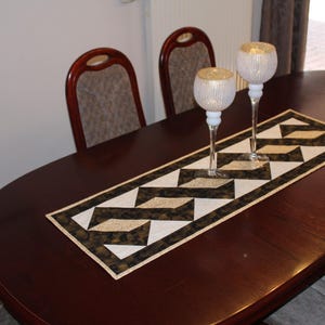 Könnte beinhalten: Ein dunkelbrauner ovaler Esstisch mit zwei passenden Stühlen. Ein geometrisch gemusterter Tischläufer in Schwarz, Weiß und Gold ist auf dem Tisch zentriert. Zwei dekorative Glaskerzenhalter mit goldenen Akzenten befinden sich auf dem Läufer.
