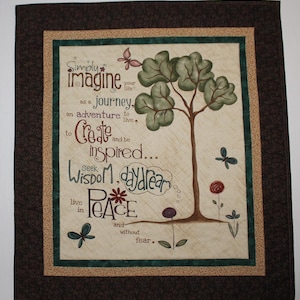 Puede incluir: Una pared colgante de tela beige con un marco marrón y un borde verde. El centro de la pared colgante presenta un árbol estilizado con hojas verdes y un tronco marrón. El texto "Simply imagine your life as a journey. An adventure to live, to create and be inspired... Seek wisdom, daydream in peace and without fear." está impreso en la pared colgante.