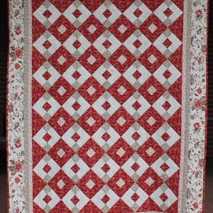 Könnte beinhalten: Eine handgefertigte Steppdecke mit einem sich wiederholenden Rautenmuster in Rot, Weiß und Beige. Die Steppdecke hat einen floralen Rand in Rot-, Beige- und Weißtönen. Die Steppdecke wird aufgehängt, um ihr Design und ihre Handwerkskunst zu präsentieren.