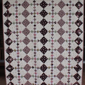 Op de afbeelding: Een quilt met een ruitpatroon in tinten roze, bruin en groen. De quilt is gemaakt van verschillende bloemenstoffen.