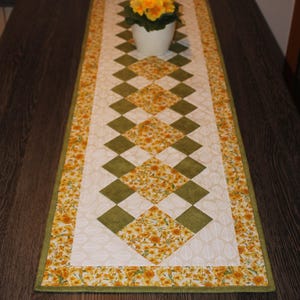 Peut inclure: Chemin de table matelassé avec un motif floral et géométrique. Le chemin de table présente un motif central de losanges verts et floraux alternés, bordé d'un imprimé floral et d'un bord vert. Un pot blanc avec des fleurs jaunes est posé dessus.