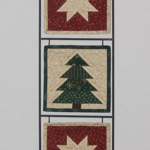 Peut inclure: Un support mural vertical en métal noir tenant trois panneaux de courtepointe carrés en tissu. Les panneaux supérieur et inférieur présentent une étoile à huit branches crème sur fond rouge foncé. Le panneau central affiche un sapin vert.