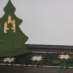 Op de afbeelding: Een groene kerstboomvormige decoratie met drie kaarsuitsparingen, verlicht met warm licht. Hij staat op een tafelloper met een patroon van groene en witte sterren. De boom is ongeveer 46 cm hoog.