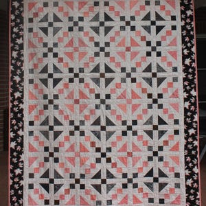Puede incluir: Una colcha rectangular con un patrón geométrico repetitivo de bloques de tela en blanco, rosa coral y gris carbón. La colcha está enmarcada por un borde ancho negro decorado con un estampado de hojas blancas y coral, con un ribete fino.