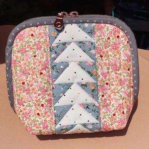 Puede incluir: Una pequeña bolsa acolchada con cremallera, de color gris y blanco, con un estampado floral. La bolsa tiene una cremallera y un tirador de cremallera decorativo.
