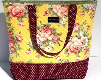 May include: A yellow quilted tote bag with a floral print and a burgundy polka dot bottom. The bag has two black straps and a label that says "100% Homemade".