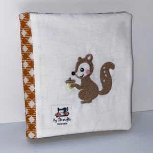 May include: A handmade, white fabric book cover with a brown and tan diamond pattern on the spine. The cover features an embroidered brown squirrel holding an acorn. The label reads "By TJ.crafts Handmade."