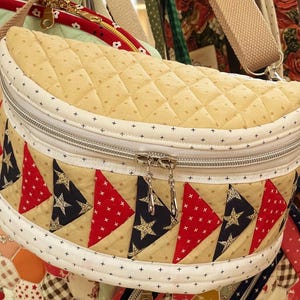 May include: A crescent-shaped quilted crossbody bag featuring a beige top panel with a diamond stitch pattern and a lower section decorated with alternating red and navy blue triangular fabric patches printed with white stars and cross patterns.