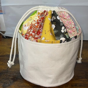 Peut inclure: Un sac à cordon blanc avec une doublure en tissu faite de tissu patchwork coloré. Le sac est en toile et a une forme ronde.