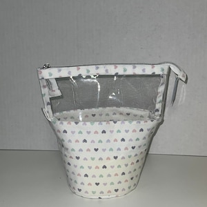 Puede incluir: Una bolsa de plástico transparente con cremallera, con un forro de tela blanca con un patrón de pequeños corazones multicolores. La bolsa está de pie sobre una superficie blanca.