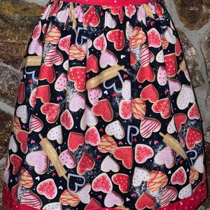 Puede incluir: Un delantal de lunares rojos y blancos con fondo negro que presenta un patrón de galletas en forma de corazón con glaseado rosa y blanco. El delantal tiene una cintura roja y un ribete rojo con lunares blancos.
