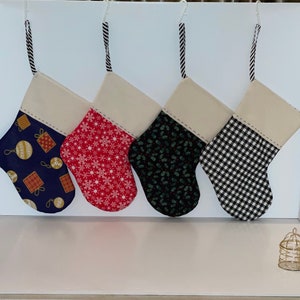 May include: Four Christmas stockings with cream-colored tops and patterned bodies. The patterns include ornaments on navy, snowflakes on red, holly on black, and a black and white gingham check. Each stocking has a striped hanging loop.