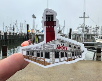 May include: A sticker depicting a red and white lighthouse and a building with the text "ANDYS". The sticker is held up in front of a harbour with boats in the background.