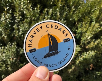 May include: A round sticker with a blue, yellow and white design. The sticker features a sailboat on a body of water with a sunset in the background. The text on the sticker reads "Harvey Cedars Est. 1894 Long Beach Island, N.J."