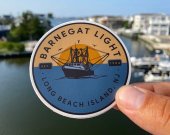 May include: A round sticker with a yellow, blue and white design. It features a ship with a sunset in the background. The text reads "Barnegat Light Est. 1904 Long Beach Island, NJ."