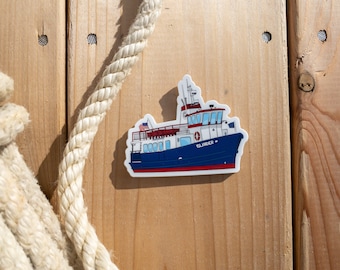 May include: A blue and white ferry boat sticker with the word "ISLANDER" on the side. The sticker is on a wooden background.