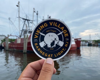 May include: A round sticker with a black background and gold lettering. The sticker says "Viking Village, Barnegat Light, NJ."  It has a white image of a fish with crossed oars and the words "Wild Caught."