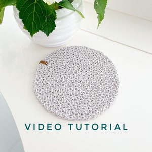 May include: A gray crocheted coaster with a brown leather loop. The coaster is on a white surface with a white vase and white flowers.