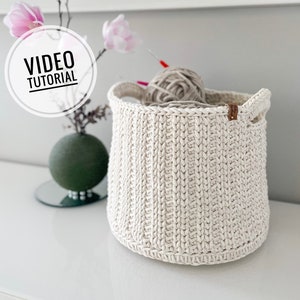 Puede incluir: Una cesta de crochet blanca con dos asas de cuero. La cesta está llena de una bola de hilo beige. El texto "VIDEO TUTORIAL" es visible en un círculo blanco.