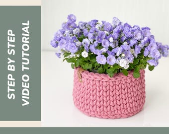 Op de afbeelding: Een roze gehaakte mand gevuld met paarse bloemen. De mand is rond en heeft een getextureerd uiterlijk. De tekst "CROCHET BASKET" staat bovenaan en "STEP BY STEP VIDEO TUTORIAL" aan de linkerkant.