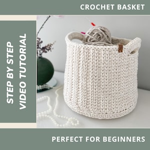 May include: A cream-colored crochet basket with handles, containing a ball of yarn and knitting needles. The text "CROCHET BASKET" is at the top, and "STEP BY STEP VIDEO TUTORIAL" is on the left. A small brown tag is attached to the basket.