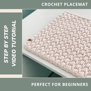 Op de afbeelding: Een beige gehaakte placemat met een getextureerd oppervlak. De afbeelding bevat bovenaan de tekst "CROCHET PLACEMAT" en links "STEP BY STEP VIDEO TUTORIAL". Een klein bruin label is aan de placemat bevestigd.