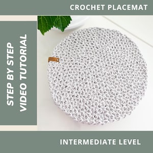Puede incluir: Un salvamanteles redondo de ganchillo gris claro con un patrón de estrella texturizado. El salvamanteles tiene una pequeña etiqueta de cuero marrón. El texto de la imagen dice "CROCHET PLACEMAT", "STEP BY STEP VIDEO TUTORIAL" y "INTERMEDIATE LEVEL".