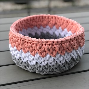 Puede incluir: Una cesta redonda de ganchillo hecha a mano en tonos coral, blanco y gris. La cesta presenta un patrón de puntada texturizada y es adecuada para almacenamiento o decoración. La combinación de colores es neutra y complementaría varios estilos de decoración del hogar.