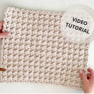 Puede incluir: Primer plano de un cuadrado de crochet beige con un patrón texturizado. El cuadrado está siendo sostenido por dos manos. El texto "VIDEO TUTORIAL" es visible en un círculo en la esquina superior derecha de la imagen.