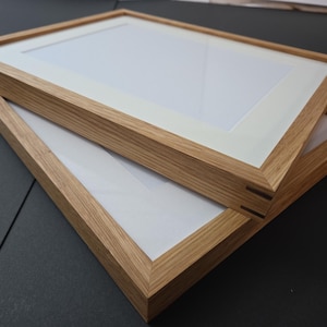 May include: Two rectangular wooden picture frames with light-colored wood grain. Each frame has a white mat and clear glass. The frames are stacked at a slight angle, showcasing the natural wood tones and clean design.