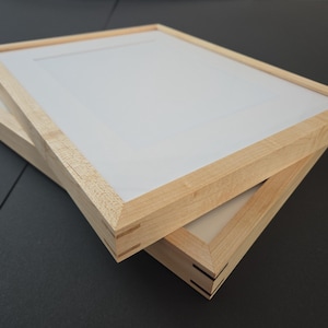 May include: Two wooden picture frames with a light-colored wood grain are stacked at a slight angle. Each frame has a white mat and a light-colored wood frame. The frames are on a dark surface.