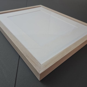 May include: A rectangular picture frame with a light wood grain finish. The frame has a white mat and a recessed area for a photograph or artwork. The frame is angled, showing the side profile and the interior.