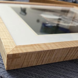 May include: Close-up of a framed artwork. The frame is made of light-colored wood with a natural grain pattern. The artwork is behind a white mat and glass. The frame's corner is visible, showing a clean, angled joint. The overall aesthetic is clean and modern.