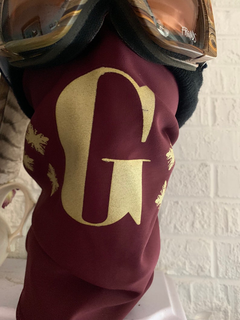 K&ouml;nnte beinhalten: Burgunderfarbener Nackenschutz mit einem goldenen Buchstaben G und goldenen Schneeflocken-Designs.