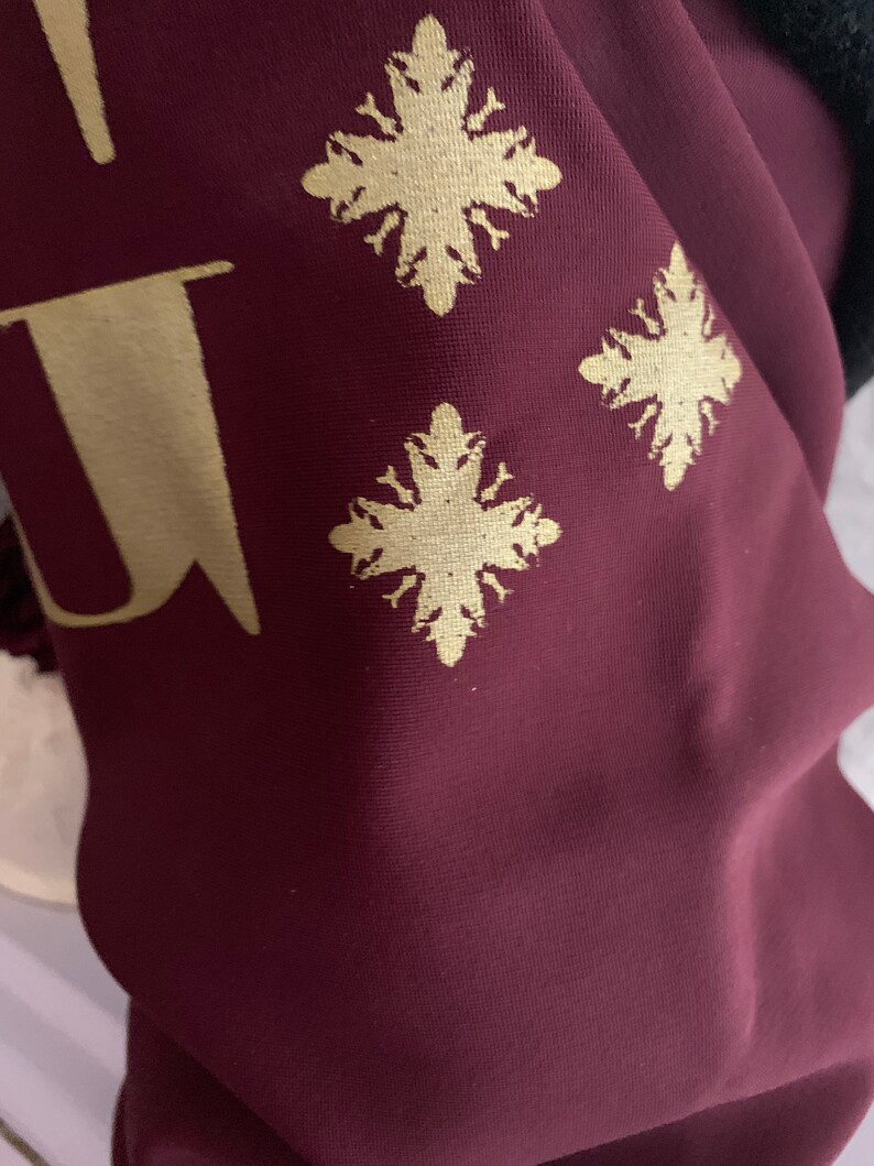 K&ouml;nnte beinhalten: Burgunderfarbener Stoff mit einem goldenen Schneeflockenmuster. Der Stoff hat eine subtile Textur.
