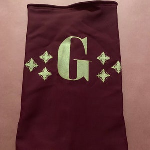K&ouml;nnte beinhalten: Ein maroonfarbener Nackentuch mit einem goldenen gestickten Buchstaben "G" und vier goldenen Schneeflocken-Designs.