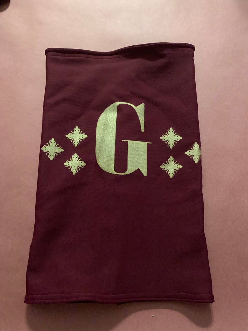 K&ouml;nnte beinhalten: Ein maroonfarbener Nackentuch mit einem goldenen gestickten Buchstaben "G" und vier goldenen Schneeflocken-Designs.