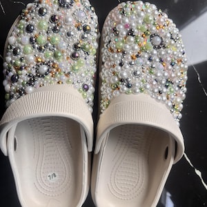 May include: A pair of cream-colored clogs, size 7/8, heavily embellished with a variety of pearls and iridescent beads. The decorative elements cover the top of the shoe, creating a glamorous, textured appearance. The clogs have a simple, functional design.