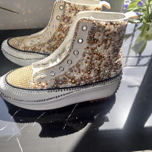 Peut inclure: Baskets montantes blanches ornées de perles dorées et nacrées. Les bouts des chaussures sont recouverts de perles dorées, et les semelles sont blanches avec une bordure en strass argentés. Les baskets ont des lacets et des œillets blancs.