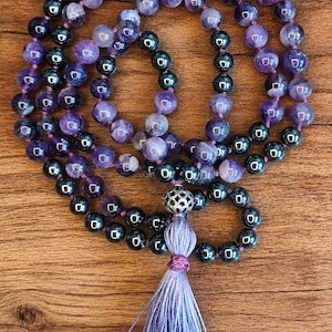May include: A beaded necklace featuring alternating purple amethyst and dark metallic hematite spheres. The strand includes a silver-toned metal bead with a cut-out pattern positioned above a light purple thread tassel at the bottom center.