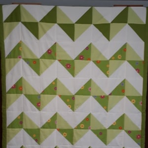 Op de afbeelding: Een groen en wit chevron-patroon quilt met een bloemenontwerp. De quilt is gemaakt van vierkante stoffen die in een geometrisch patroon aan elkaar zijn genaaid.