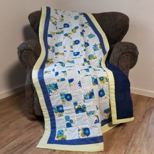Peut inclure: Une courtepointe en patchwork avec un motif floral bleu, blanc et jaune. La courtepointe est pliée et drapée sur un fauteuil marron.
