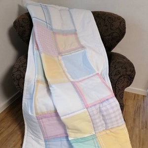 Op de afbeelding: Een patchworkdeken gemaakt van vierkanten van verschillende gekleurde stoffen. De deken is gemaakt van verschillende pastelkleuren, waaronder roze, blauw, geel en groen. De deken is gevouwen en over een stoel gelegd.