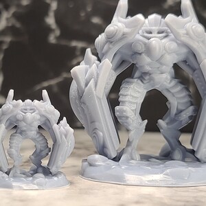 May include: Two gray, detailed miniature figurines of a fantasy character with large wings and armor, next to a blue, multi-sided die. The figurines are on a light surface with a dark, marble-like background.