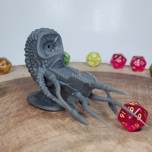 May include: A gray 3D printed miniature of a creature with a large eye and multiple tentacles. The creature is sitting on a wooden surface with several colorful dice around it.