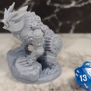 May include: A gray 3D printed miniature of a creature with owl-like features and a mechanical design. The creature is standing on a rock base and is about the size of a standard dice.